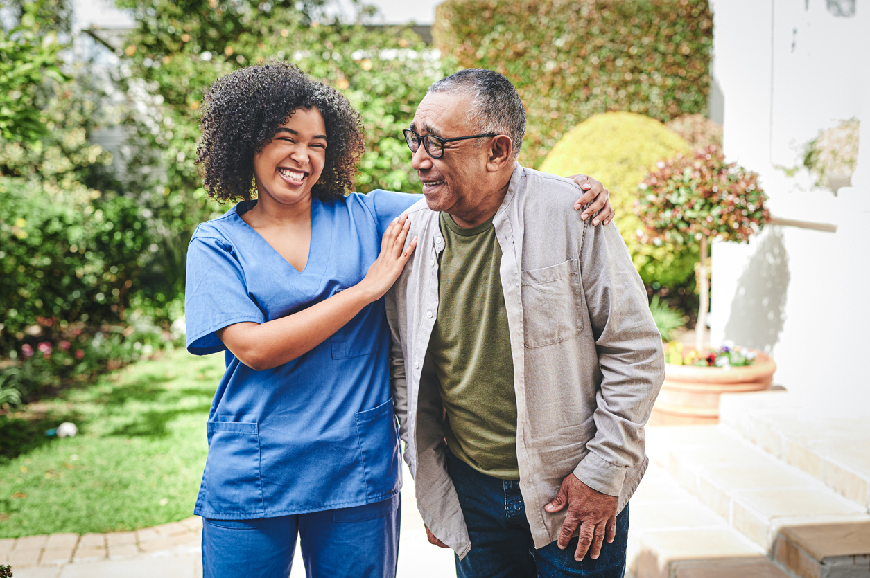 An elderly man laughing with a young female caregiver in blue scrubs outdoors